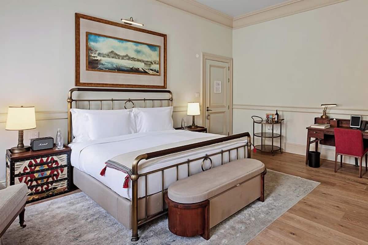 A hotel room with a bed, bedside table, lamp, desk, chair, and a painting on the wall.