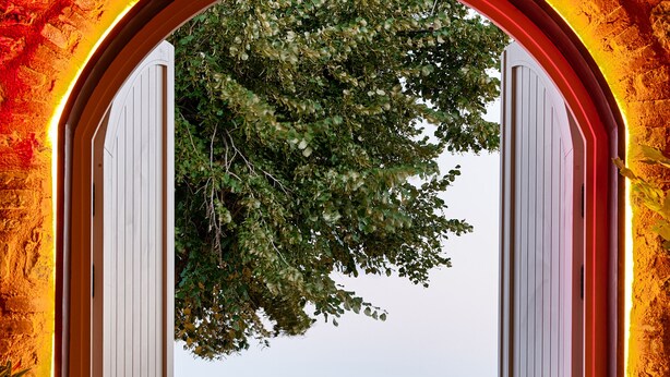 Uma porta aberta dá para um terraço com vista para o mar, uma cadeira de madeira e uma mesa.