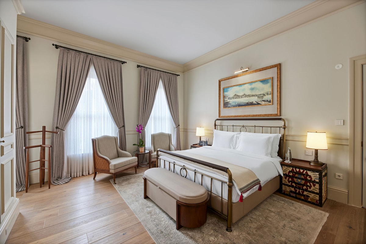A hotel room with a large bed, two bedside tables with lamps, a chair, a bench, and a painting on the wall.