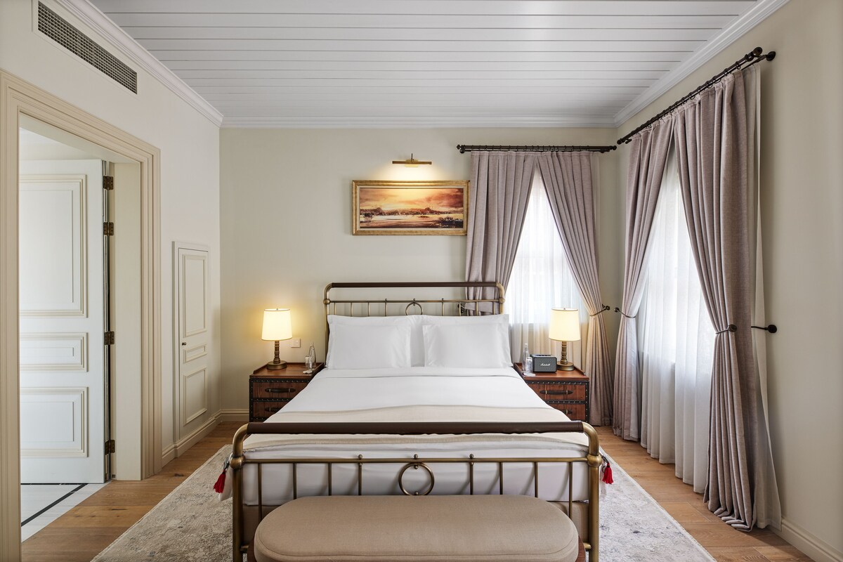 A bedroom with a bed, two bedside tables, a nightstand, a window with curtains, a painting on the wall, and a door.