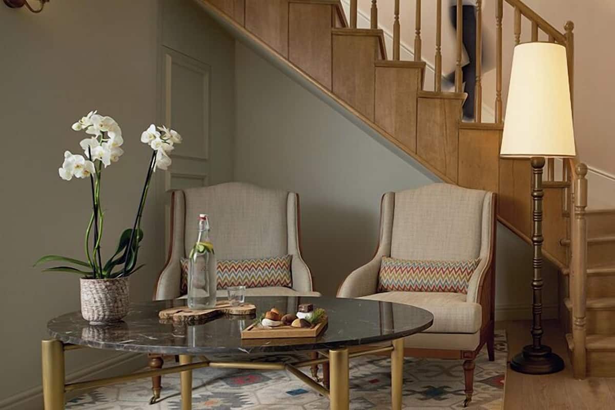 A staircase with wooden steps and a handrail, a round table with a glass top, two upholstered chairs with patterned cushions, and a floor lamp.
