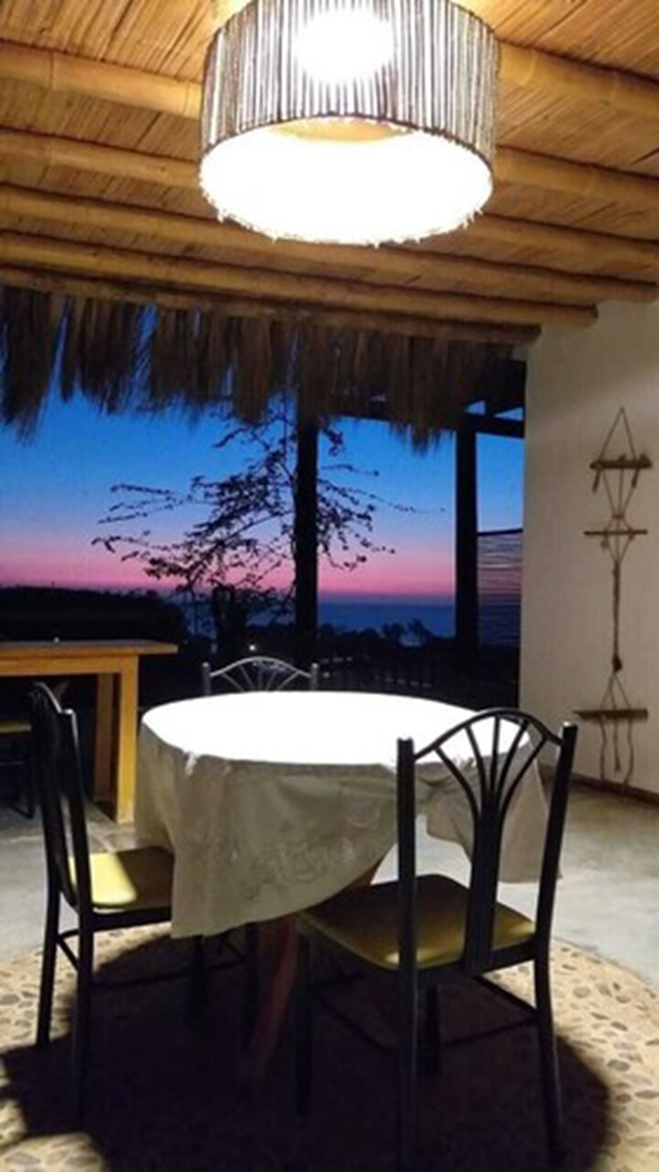 Un comedor con una mesa redonda y sillas, un techo de paja y vistas al atardecer.