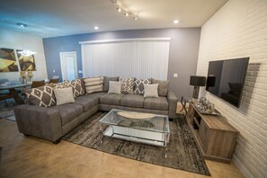 Un salon moderne avec un canapé d’angle, une table basse en verre et un téléviseur à écran plat fixé à un mur en briques.