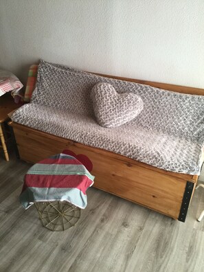 En træsofa med gråt, struktureret betræk og en pude i hjerteform. En lille, rund skammel med en stribet pude står foran sofaen.