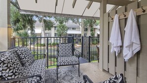 Un balcón con un sofá estampado, una silla y una mesita. Se ven toallas colgadas en un tendedero y una vista de zona residencial con árboles y edificios.