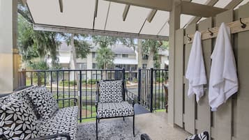 Un balcon avec un canapé à motifs, une chaise et une petite table. On y voit des serviettes suspendues à un étendoir, ainsi qu’une vue sur un quartier résidentiel avec des arbres et des immeubles.