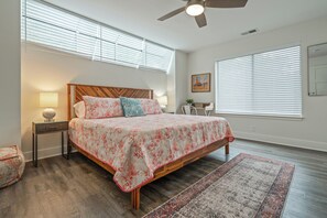 Une chambre à coucher comprenant un lit, une table de chevet, une lampe, un ventilateur de plafond et une grande fenêtre avec des stores.