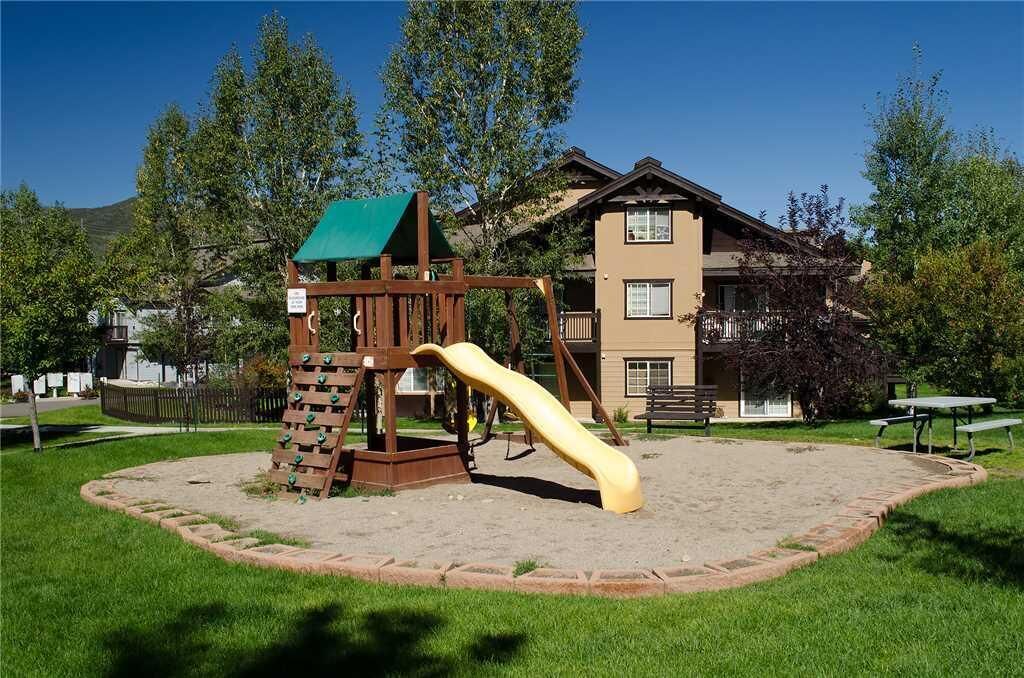 A playground with a slide and climbing structure, surrounded by a grassy area and a building in the background.