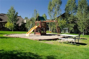 Children can play on the playground equipment, including a slide, picnic table, and bench, surrounded by a well-maintained lawn.