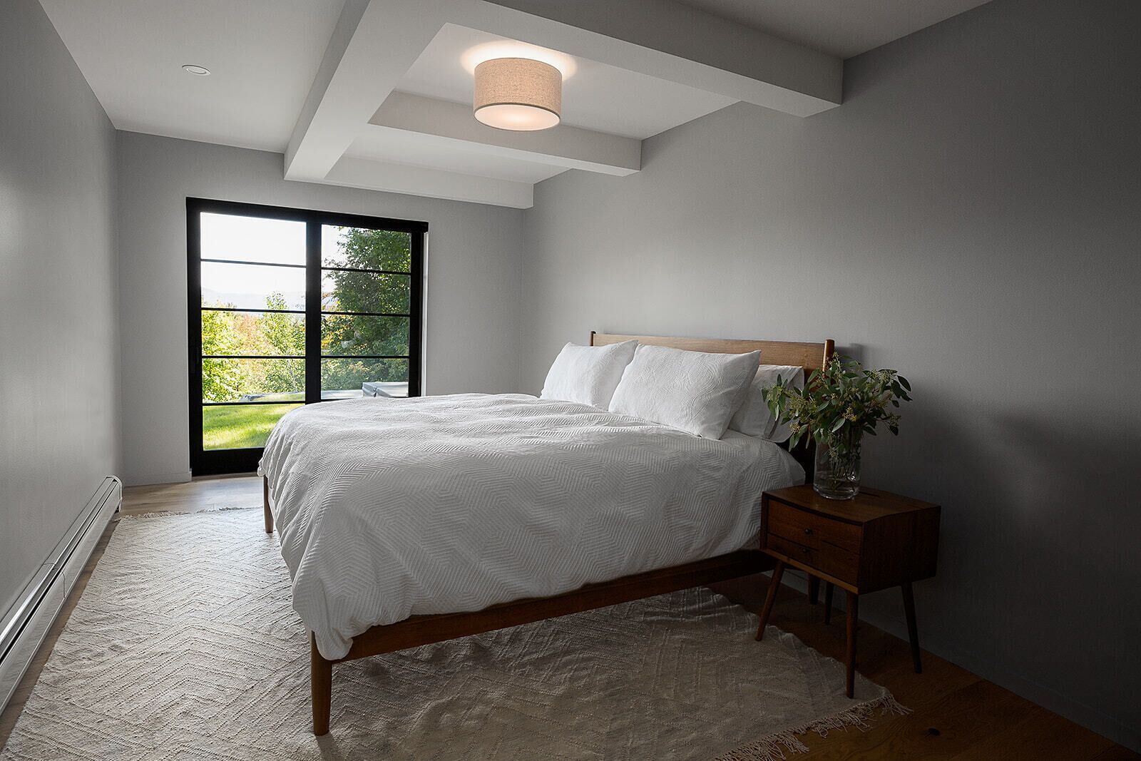 A bedroom with a large window, a bed with white bedding, a wooden headboard, a bedside table with a vase, and a rug.