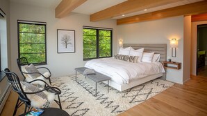 A modern bedroom with a large bed, two windows with a view of trees, a chair, a bench, and a nightstand with a lamp.