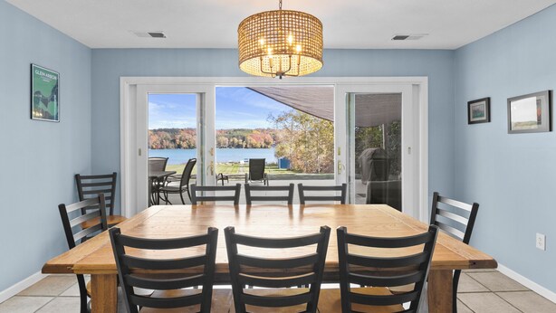 A dining area with a large wooden table and chairs, a chandelier, and a view of a lake and trees through glass doors. - Pet friendly, waterfront, home on Lake Leelanau. Pontoon available for rent (Lake Leelanau)