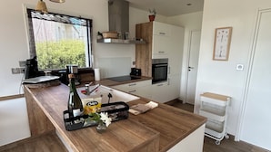 Een moderne keuken met een centraal eiland, houten aanrechtbladen, hangende pendelverlichting en uitzicht op groen.