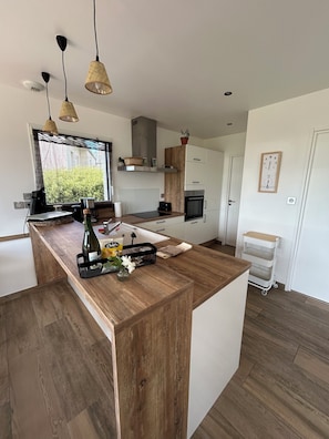 A modern kitchen with a central island, wooden countertops, hanging pendant lights, and a view of greenery outside. - Villa in Roscoff with heated indoor pool (Roscoff)
