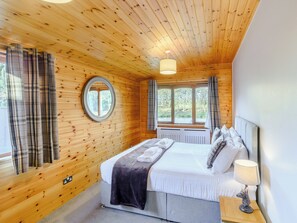 A wooden-paneled room with a bed, a round mirror, a window with curtains, and a nightstand with a lamp. - 4 bedroom accommodation in Cayton, near Scarborough (Cayton, near Scarborough)