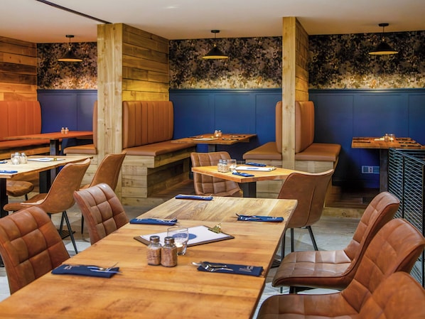 Un restaurant avec des tables et des banquettes en bois, des sièges en cuir et des murs bleus ornés d’un papier peint à motifs floraux.