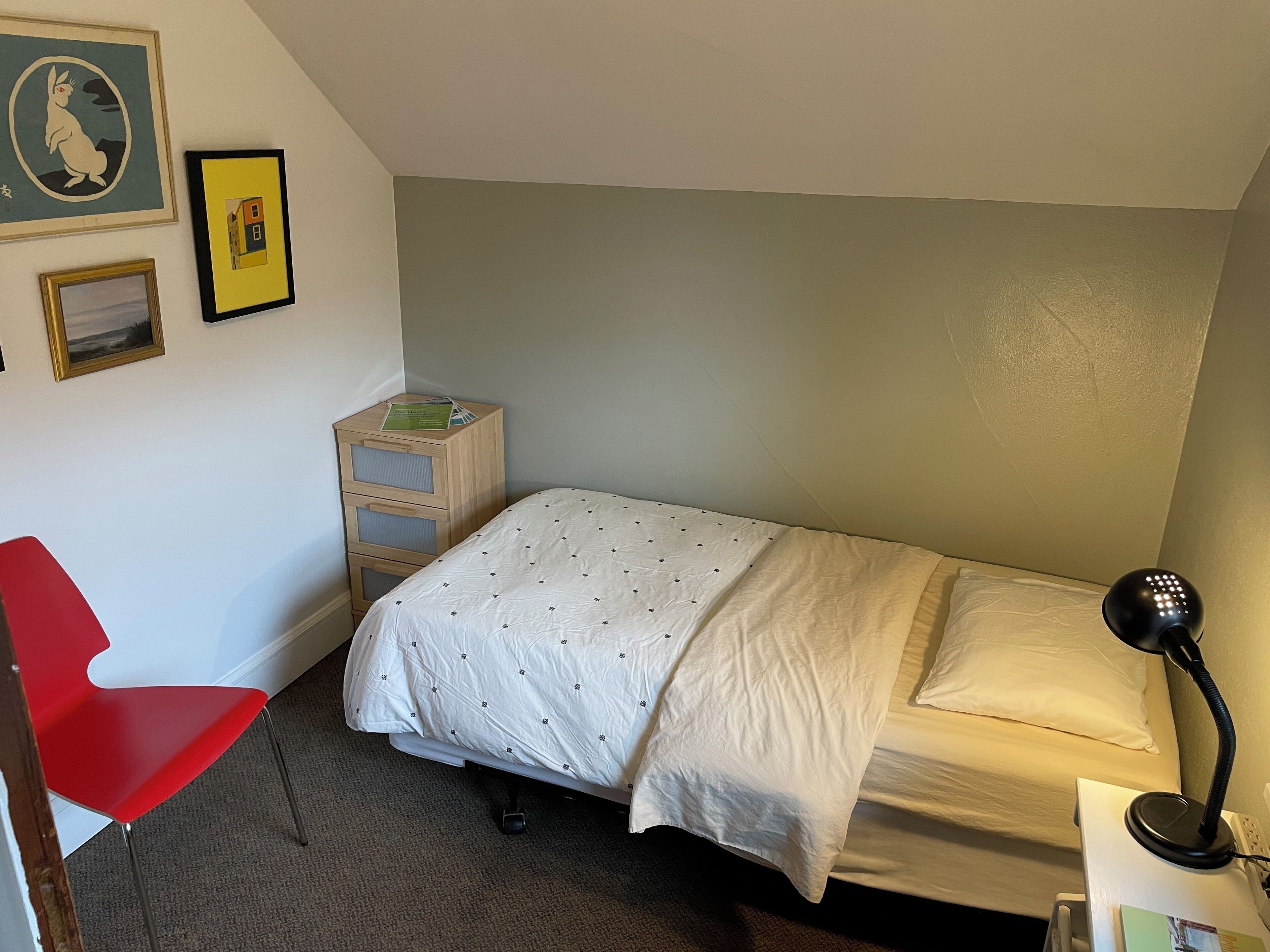 Ett litet, välordnat sovrum med en enkel säng, en röd stol, ett sängbord med en lampa och ett litet träskåp.