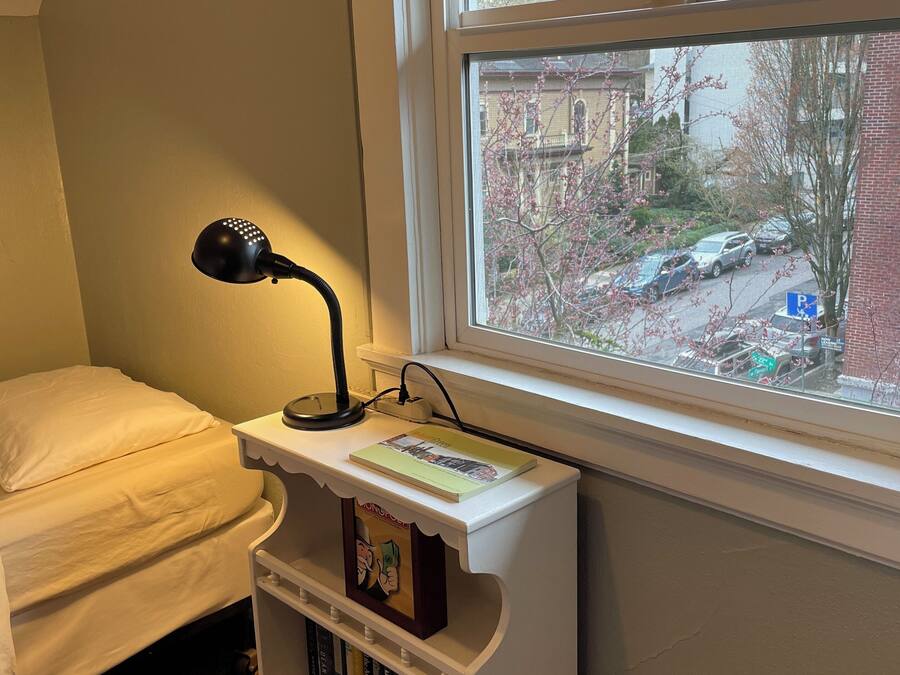 Een slaapkamer met een enkel bed, een nachtkastje met een lamp, een klein boekenrek en een raam met uitzicht op de straat en bomen.