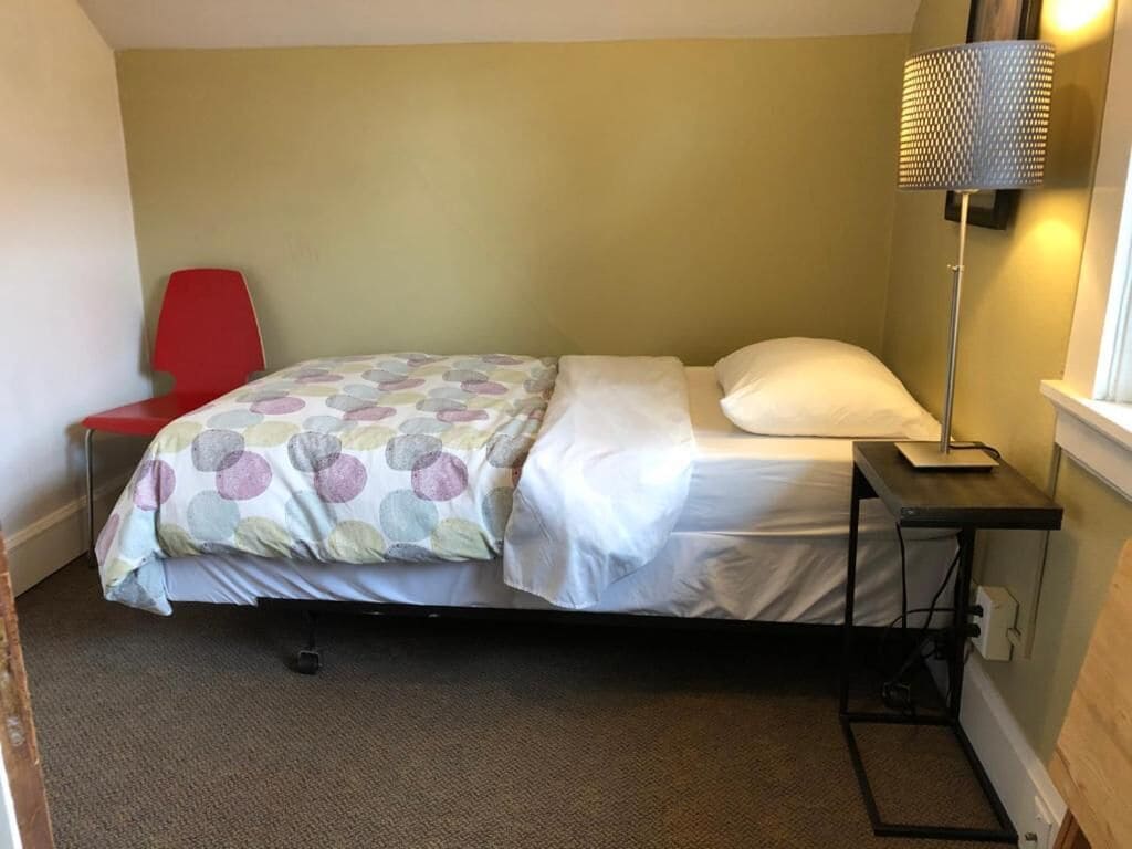 A small, single-bed room with a red chair, a bedside table with a lamp, and a window.