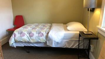 A small, single-bed room with a red chair, a bedside table with a lamp, and a window.