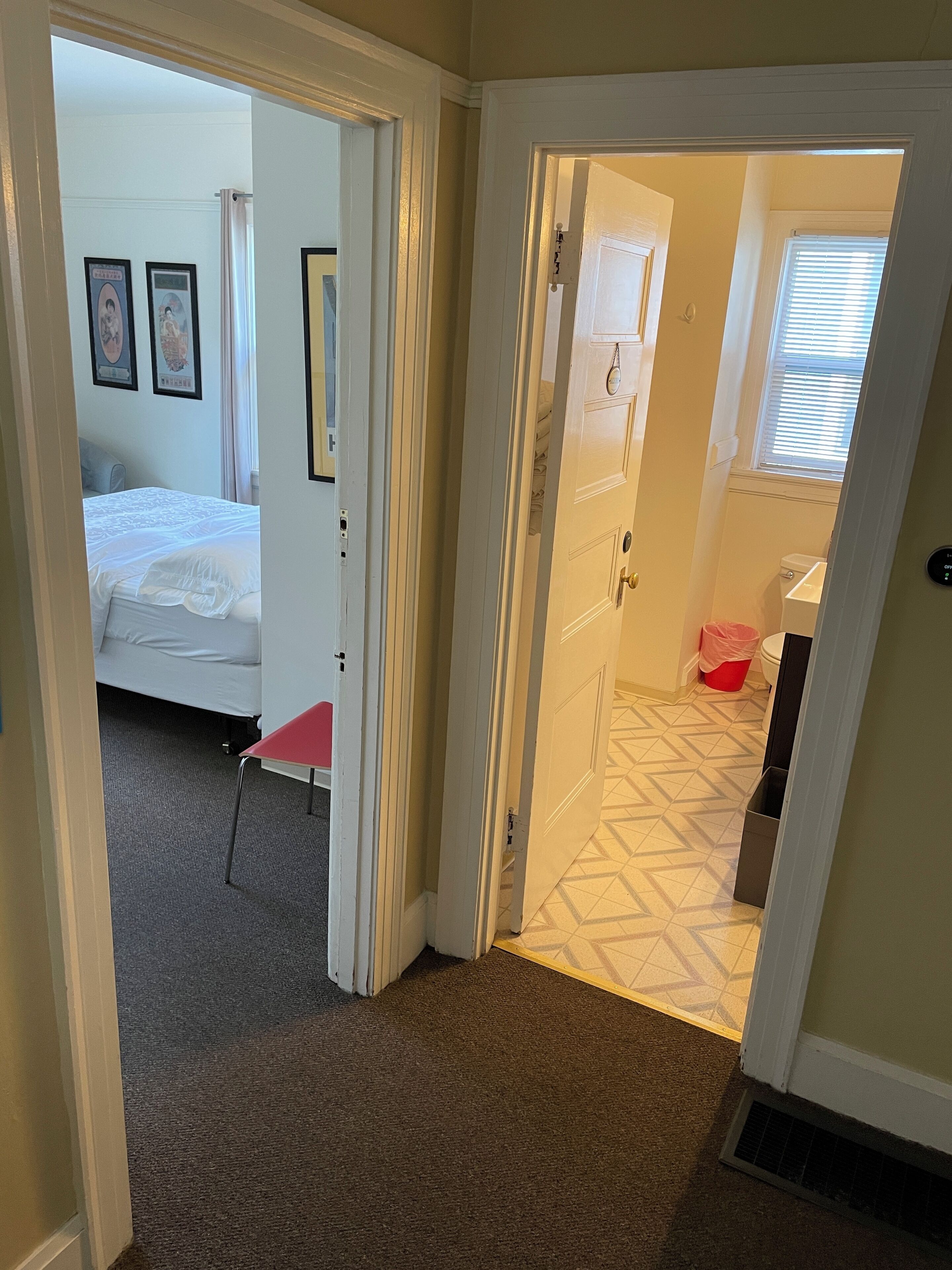 En hall som leder till ett sovrum med en säng, en röd stol och ett badrum med toalett och handfat.