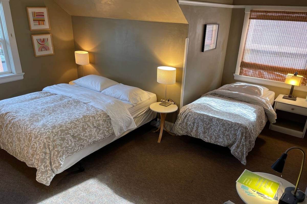 Une chambre d’hôtel avec deux lits, une petite table ronde, un bureau avec une lampe et une fenêtre avec des stores.