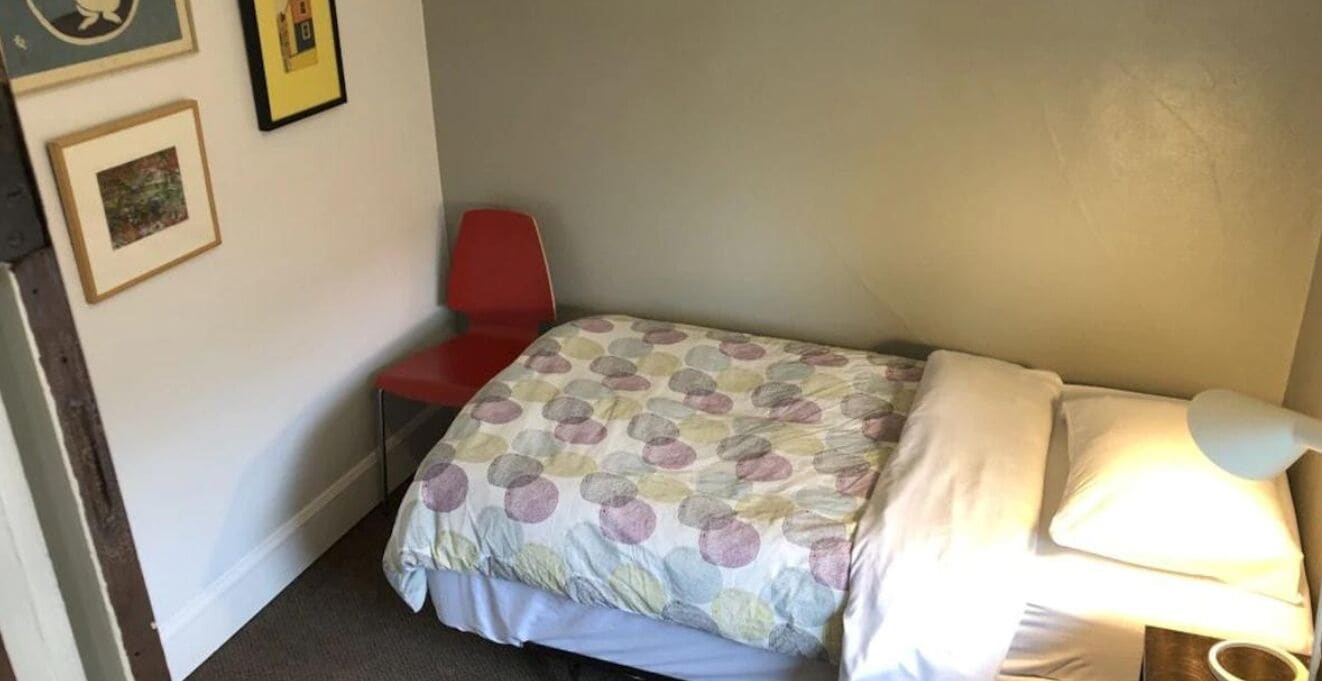 Ein kleines Zimmer mit einem Einzelbett, einem roten Sessel, einem Nachttisch mit Lampe und gerahmten Bildern an der Wand.