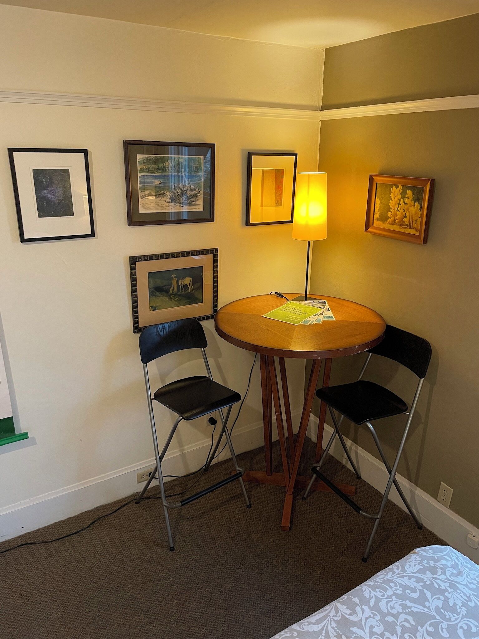 A small room with a round wooden table, two foldable chairs, a lamp, and several framed pictures on the wall.