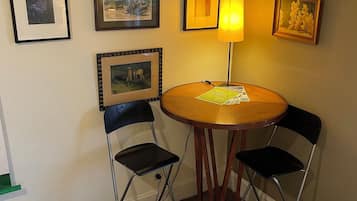 A small room with a round wooden table, two foldable chairs, a lamp, and several framed pictures on the wall.