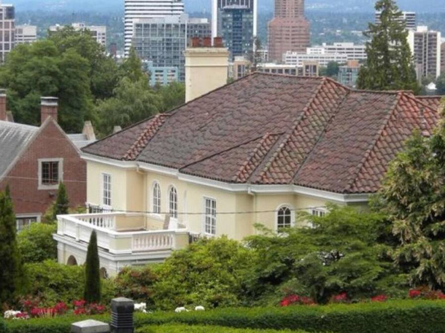 A historic building with a tiled roof, surrounded by well-maintained hedges and a cityscape in the background.