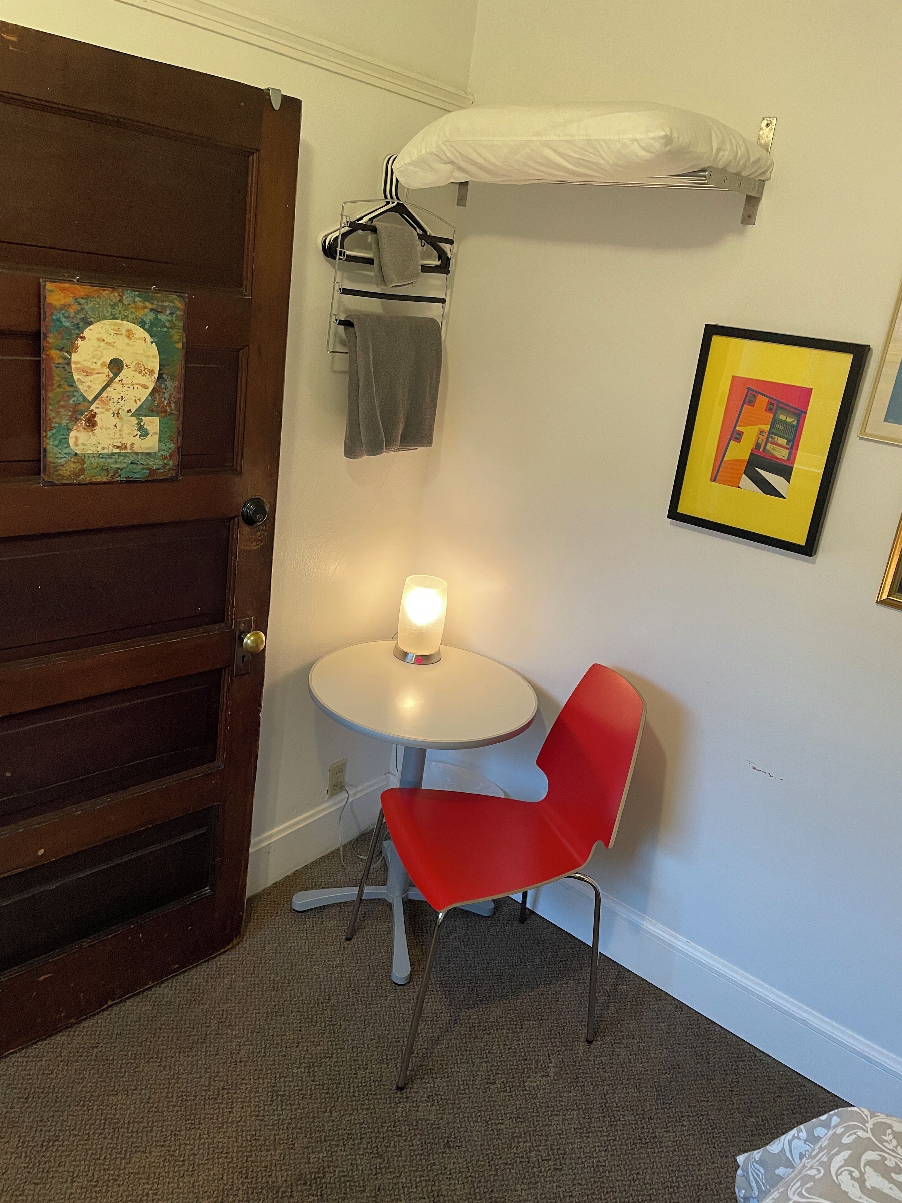 A room with a wooden door, a round table with a red chair, a hanging towel, and a framed picture on the wall.