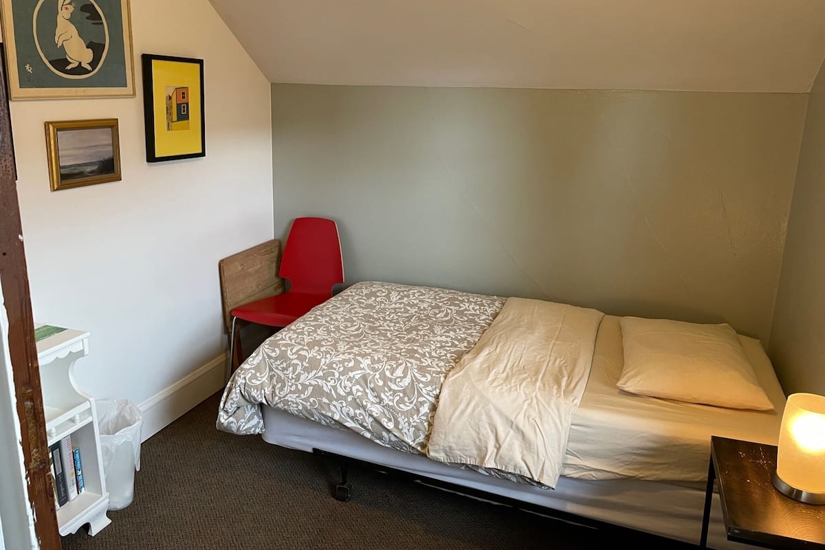 Une petite chambre avec un lit simple, une chaise rouge, une table de chevet avec une lampe et deux tableaux encadrés au mur.