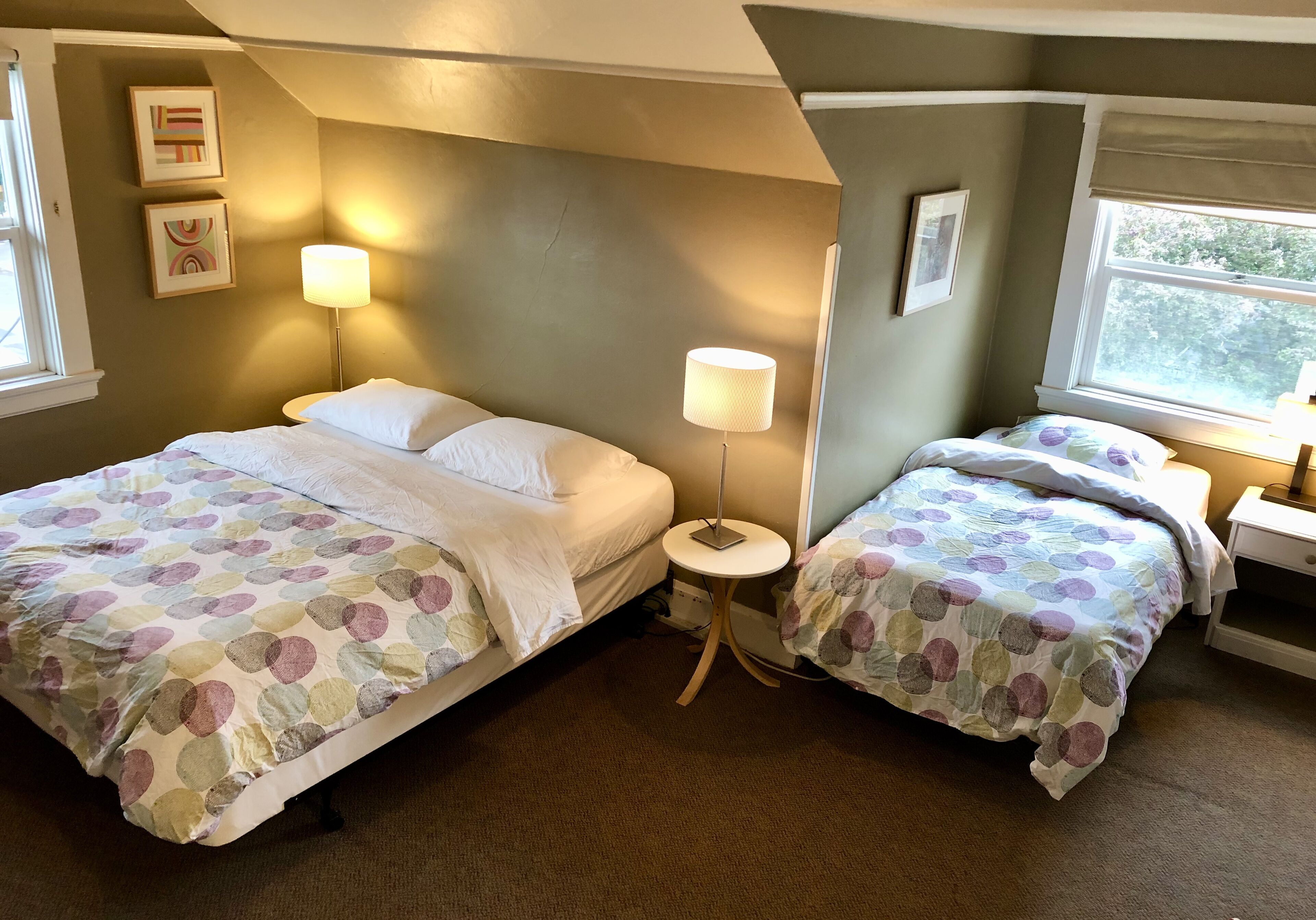 A hotel room with two beds, each with a polka dot bedspread, a small round table with a lamp, and a window with a view of greenery.