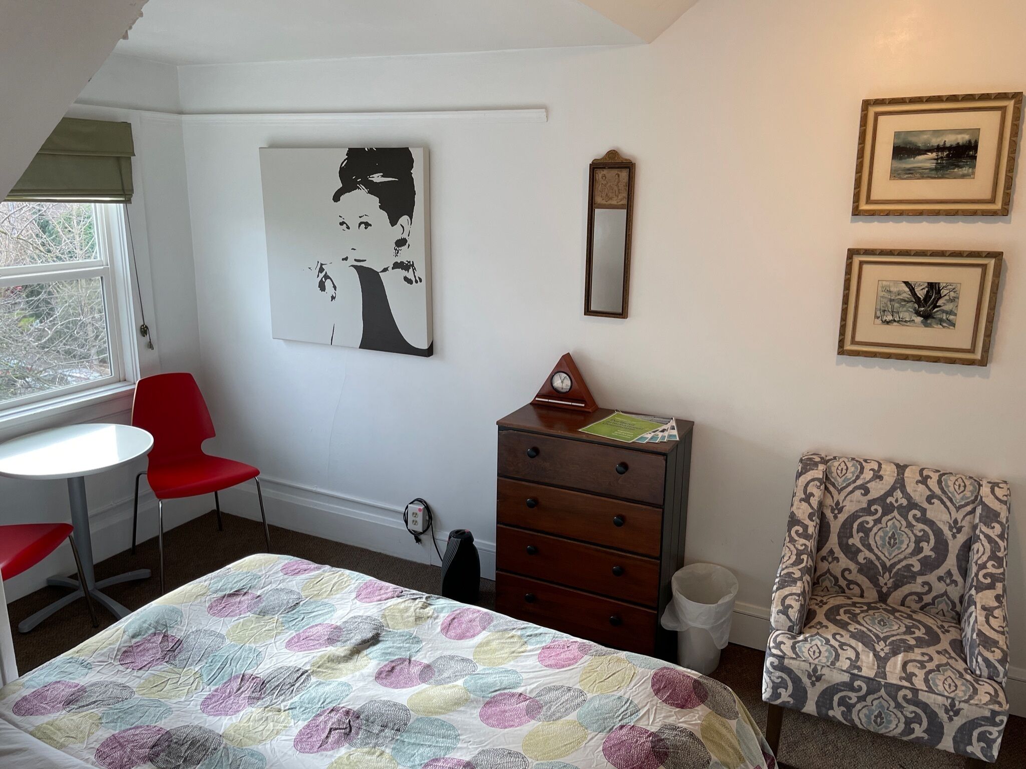 A bedroom with a bed, a red chair, a dresser, and a patterned armchair. There is a window with a view of trees, a painting on the wall, and a clock.