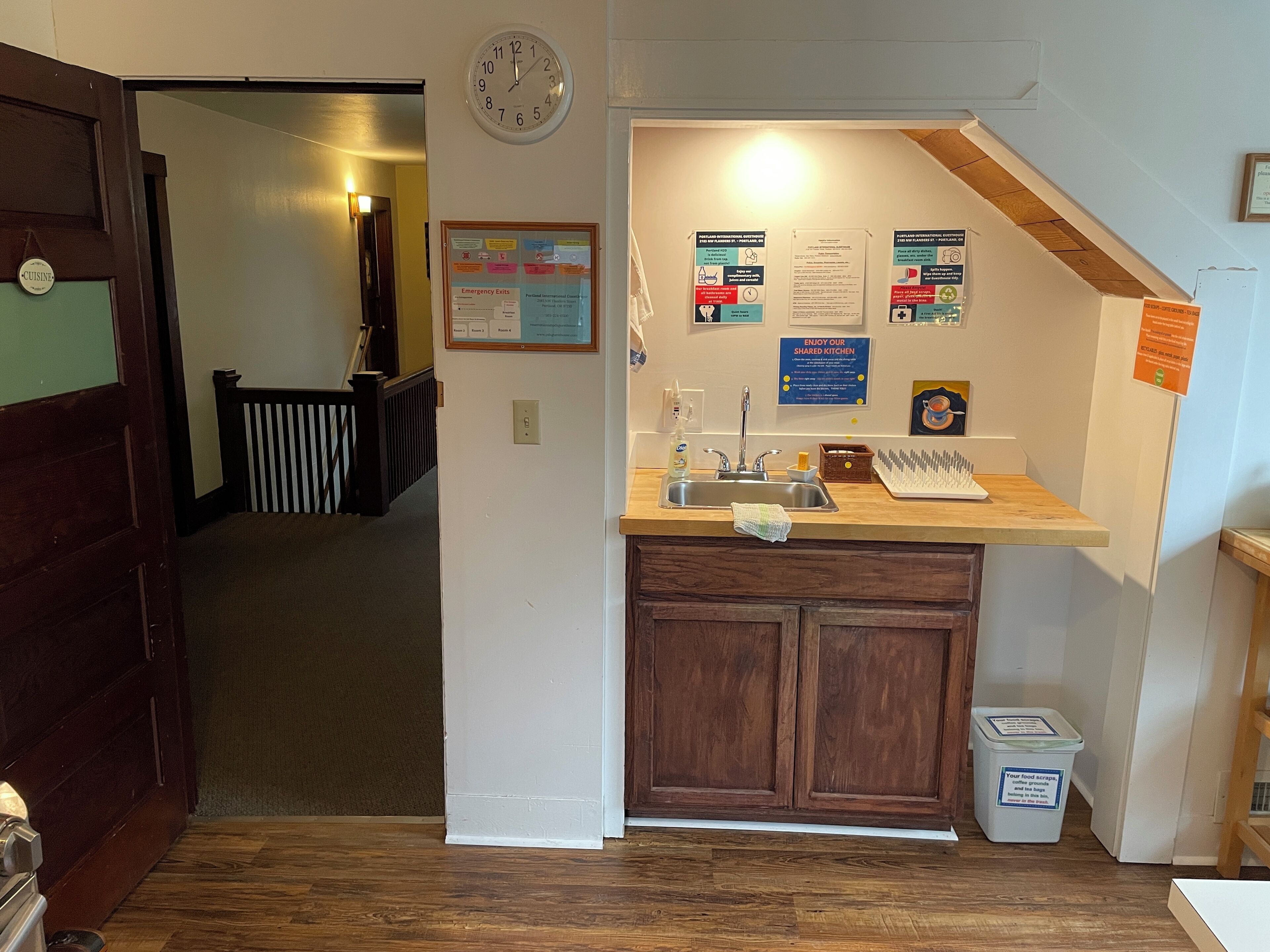 A kitchen area with a sink, countertop, and cabinets. There are multiple signs and notices on the wall, including one about emergency exits. A clock is visible on the wall, and a trash can is placed near the sink.
