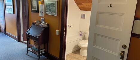 A hallway with a wooden desk and framed pictures on the wall, leading to a bathroom with a toilet and a shower area.