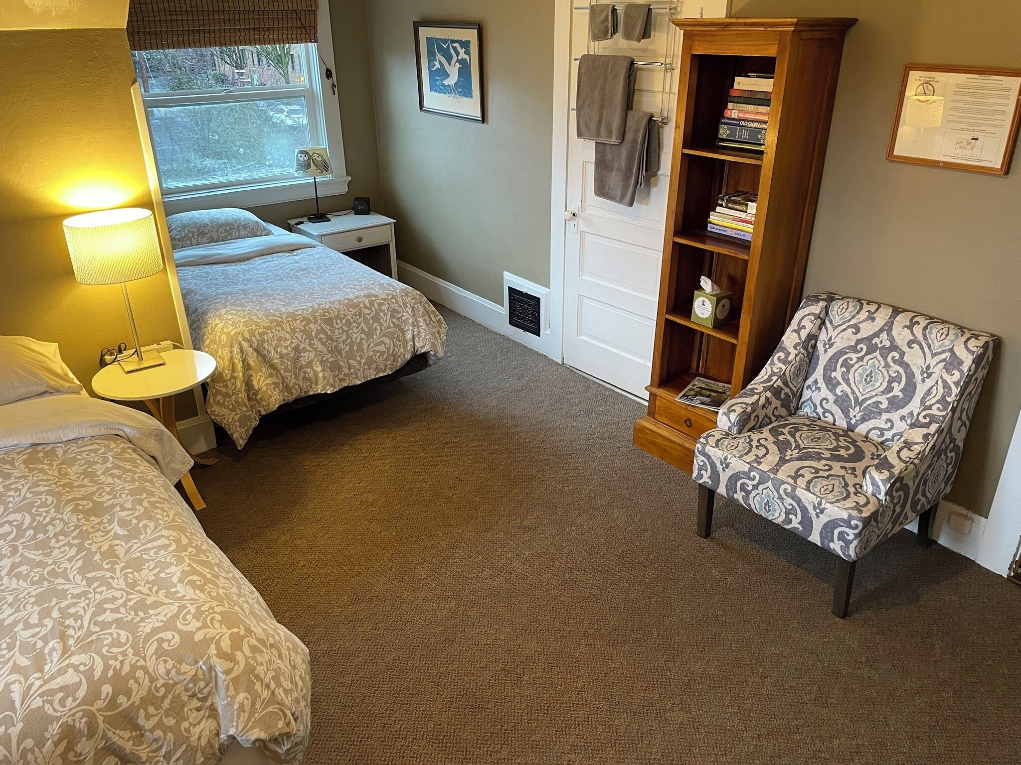 A hotel room with two beds, a bedside table with a lamp, a chair, a bookshelf, and a window with blinds.