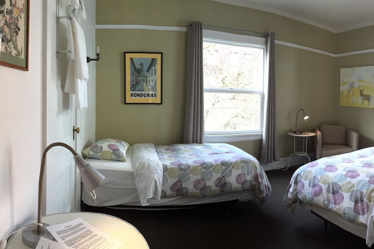 Une chambre d’hôtel avec deux lits, une petite table, une chaise, une lampe et une fenêtre avec des rideaux.