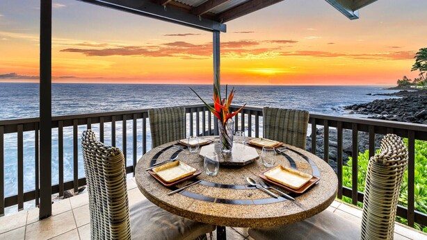 A round table with a glass centerpiece, surrounded by wicker chairs, overlooking a sunset over the ocean.