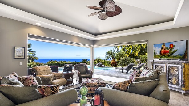A spacious living room with a large sofa, armchairs, and a coffee table. There is a ceiling fan, a television, and a view of the ocean through large windows.