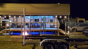 Un grand camping-car doté d’une terrasse couverte et de nombreuses fenêtres, entouré d’une clôture et avec une remorque devant.