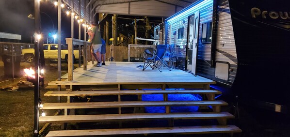 A covered outdoor area with wooden steps leading to a deck, a trailer, and a few chairs. - Fishing Camper in Shell Beach Louisiana (St Bernard)