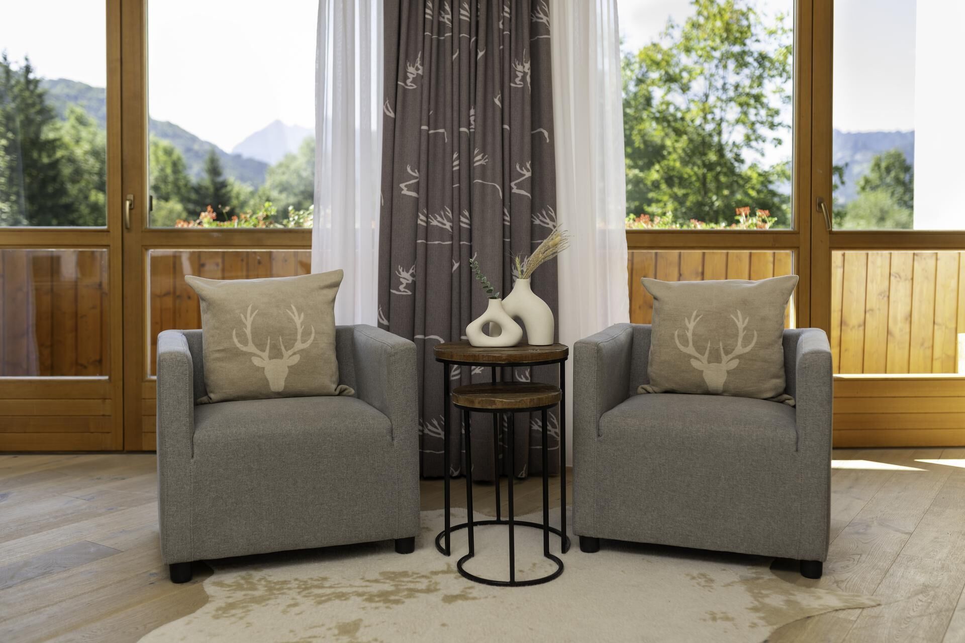 Two grey armchairs with deer head cushions, a small round table with two white vases, and a view of mountains through large windows.