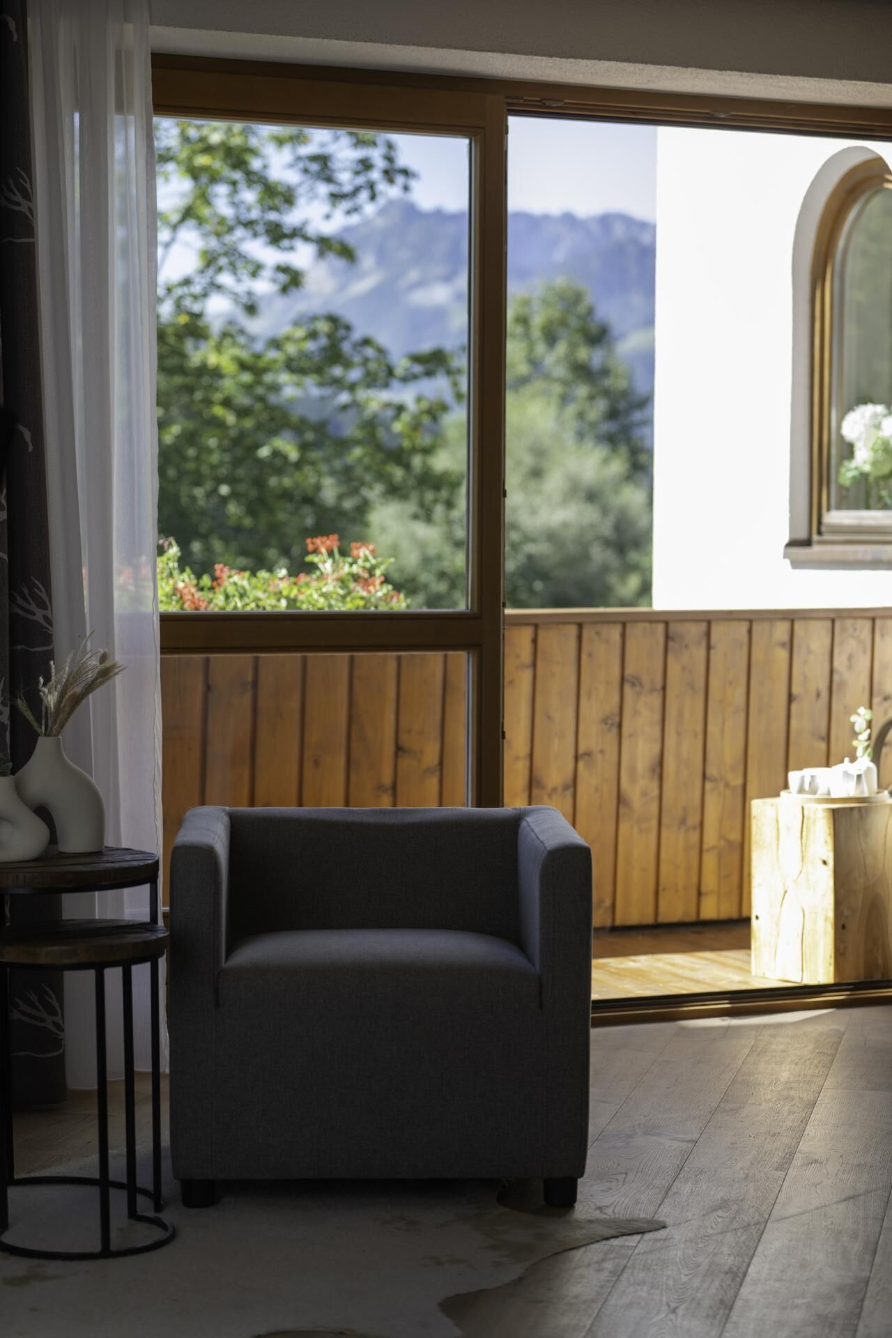 Una habitación con un ventanal grande, un sofá gris, una mesita redonda con un florero y una terraza de madera afuera.