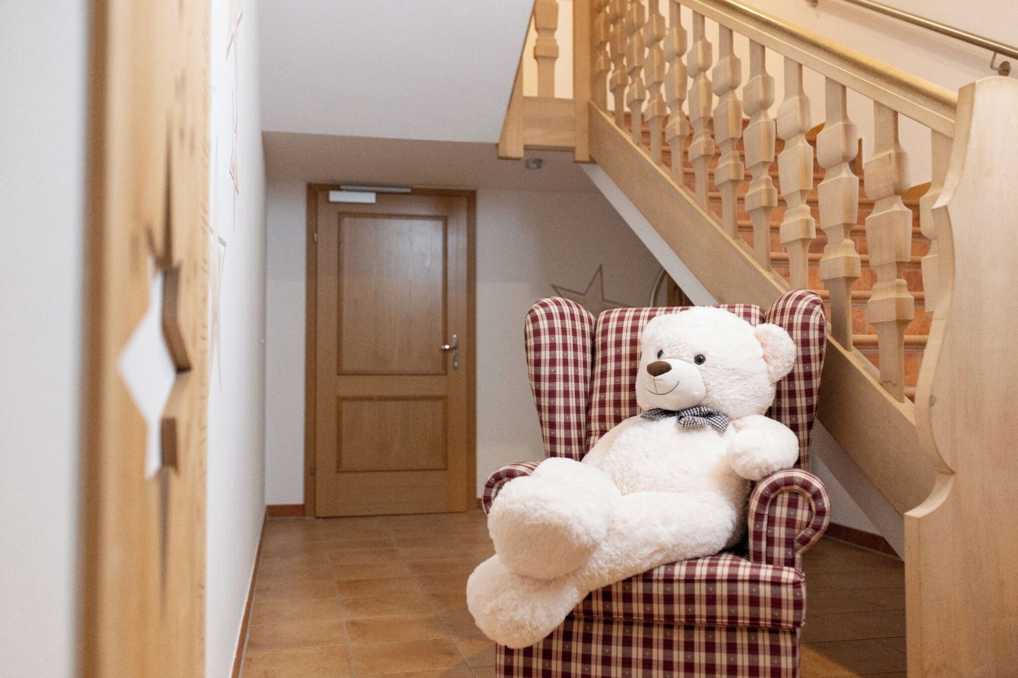 Un osito de peluche blanco y mullido sentado en un sillón a cuadros en un pasillo con una escalera de madera.