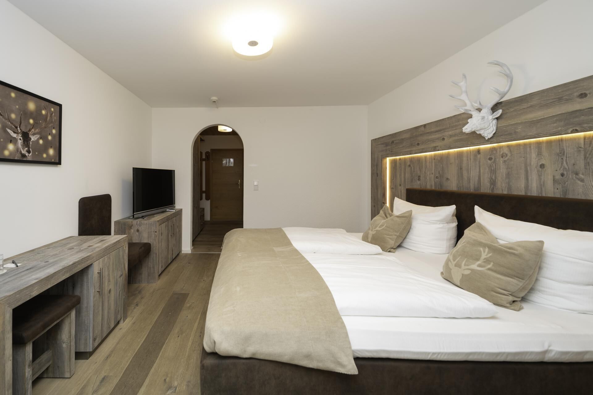 Une chambre d’hôtel moderne dotée d’une tête de lit en bois, d’un téléviseur à écran plat, d’un bureau et d’un lit avec du linge de lit blanc et des coussins décoratifs.