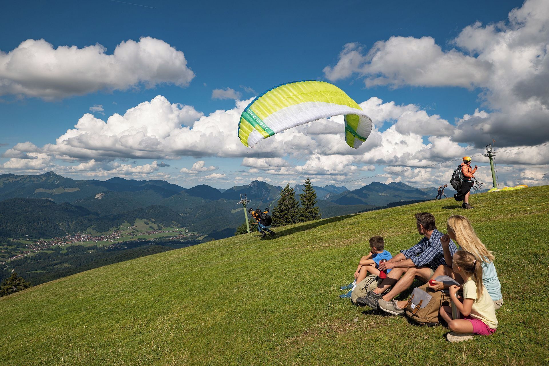 Eine Gruppe von Menschen auf einer grünen Anhöhe mit Blick auf Berge und eine Stadt darunter.