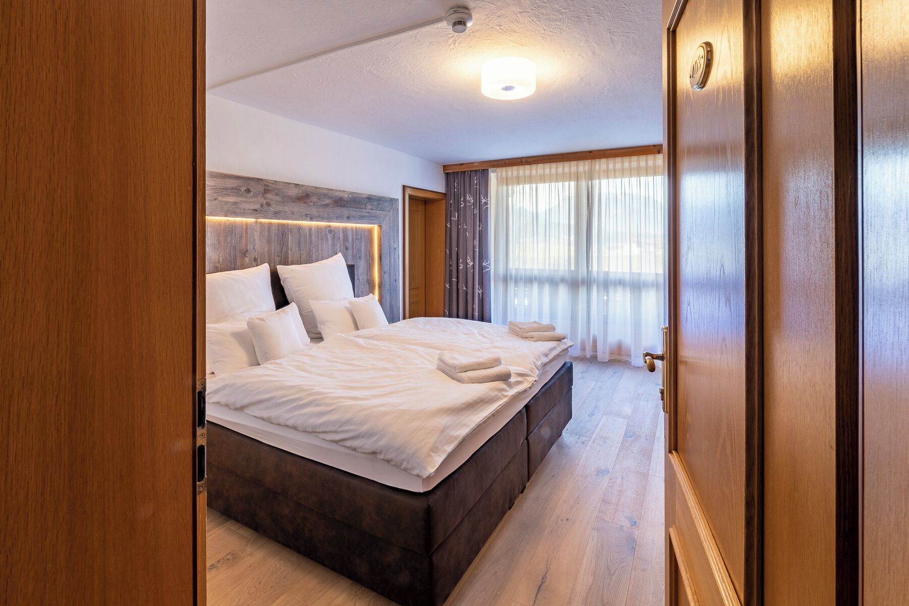 A hotel room with a large bed, wooden headboard, and a view of the outdoors through sheer curtains.