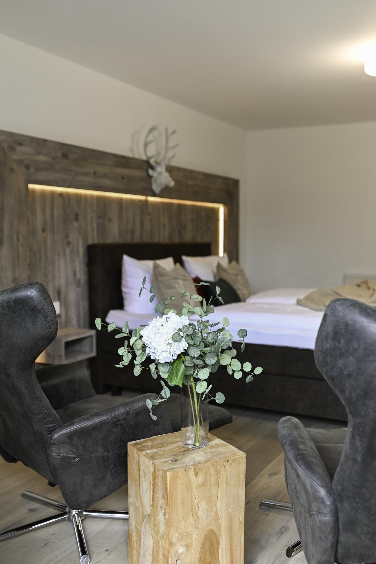 Ein modernes Schlafzimmer mit einem Holzkopfende, einem Bett mit weißer Bettwäsche und einer Vase mit weißen Blumen auf einem Holzschemel.