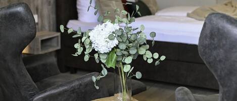 Ein modernes Schlafzimmer mit einem Holzkopfende, einem Bett mit weißer Bettwäsche und einer Vase mit weißen Blumen auf einem Holzschemel.